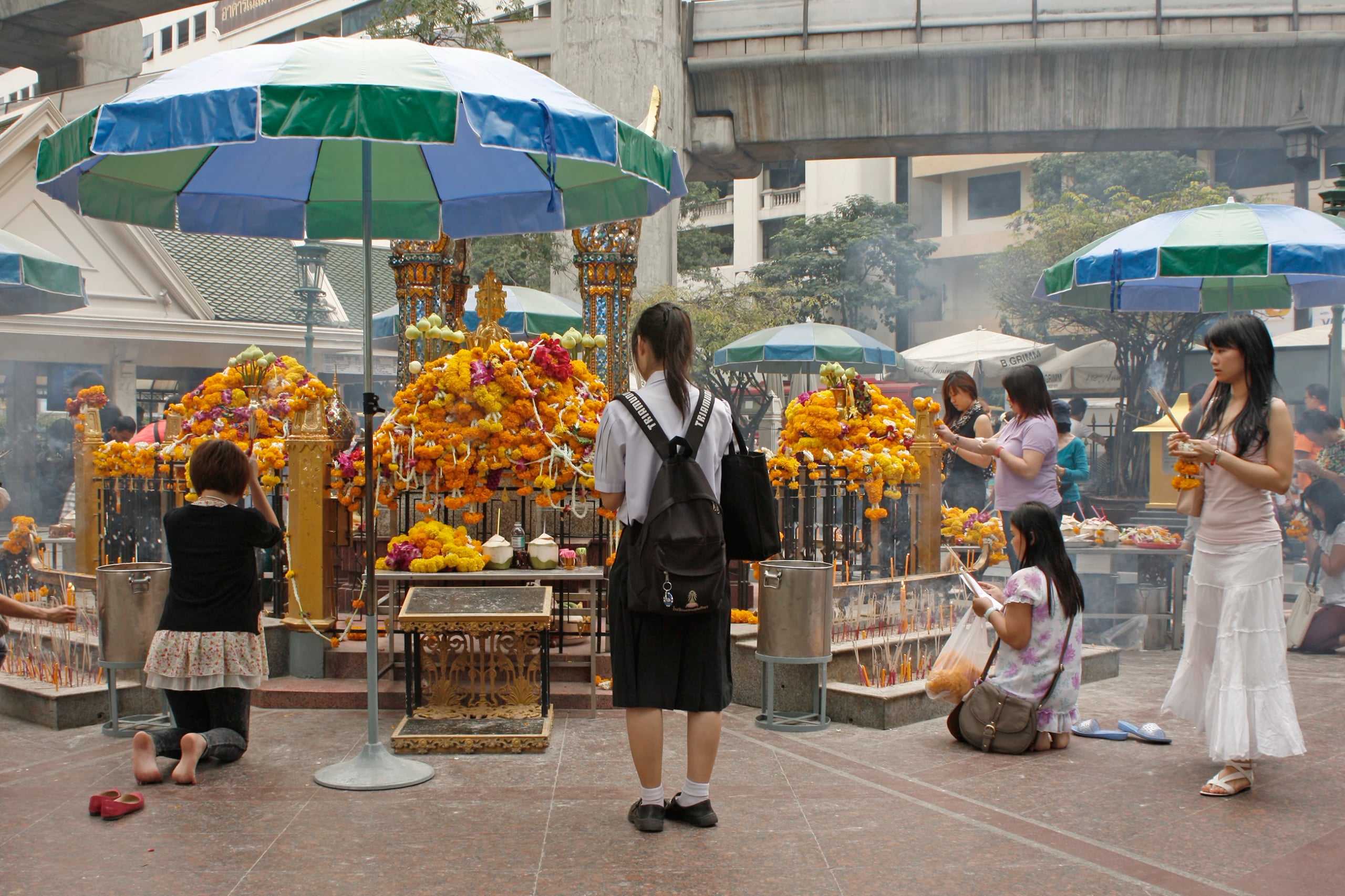 Worshippers kneeling and lighting incense at a street shrine, marigold garlands heaped on the altar under striped umbrellas