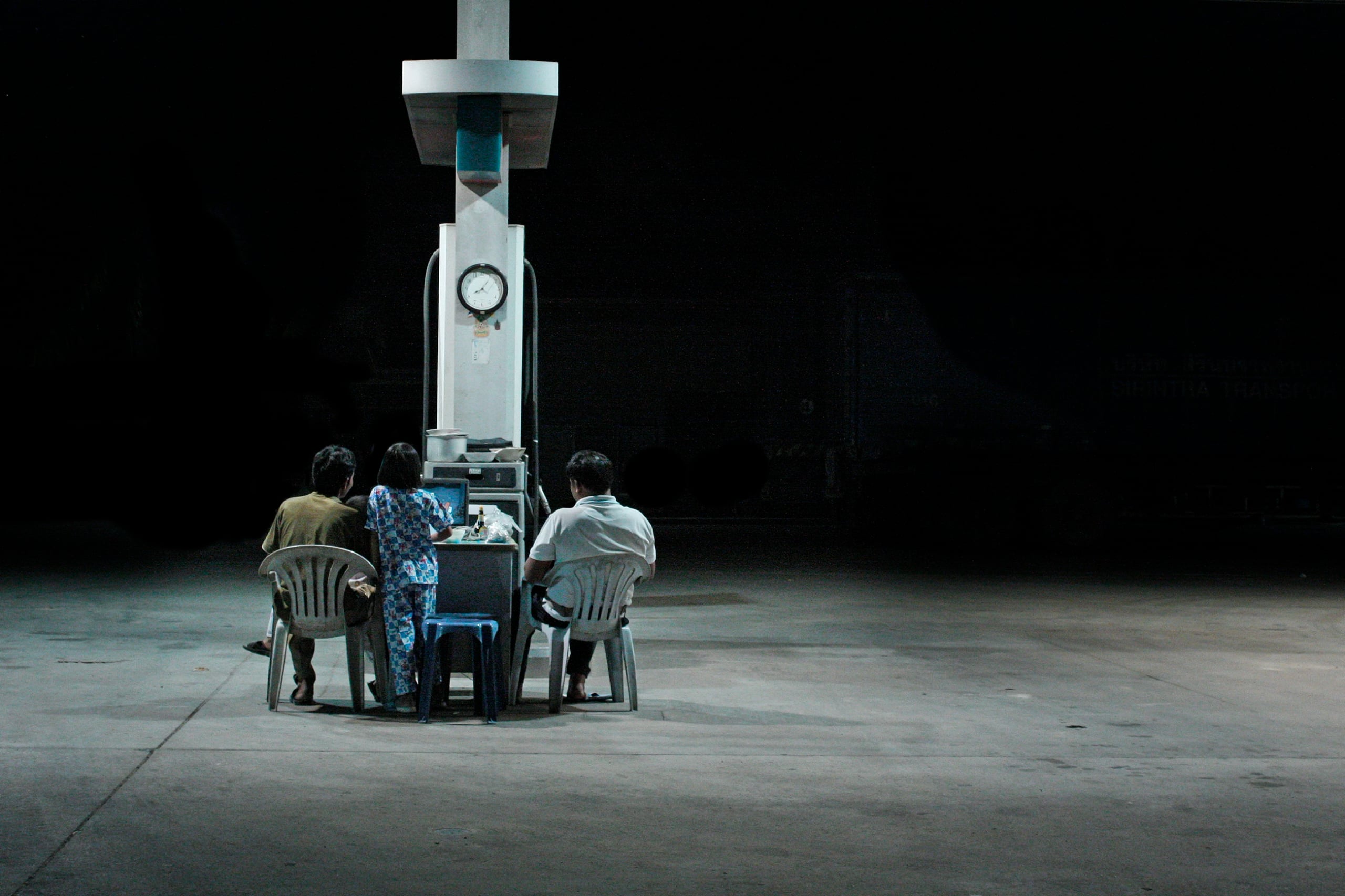 Three people on plastic chairs watching a small television at the foot of a lit petrol pump, the forecourt dark around them