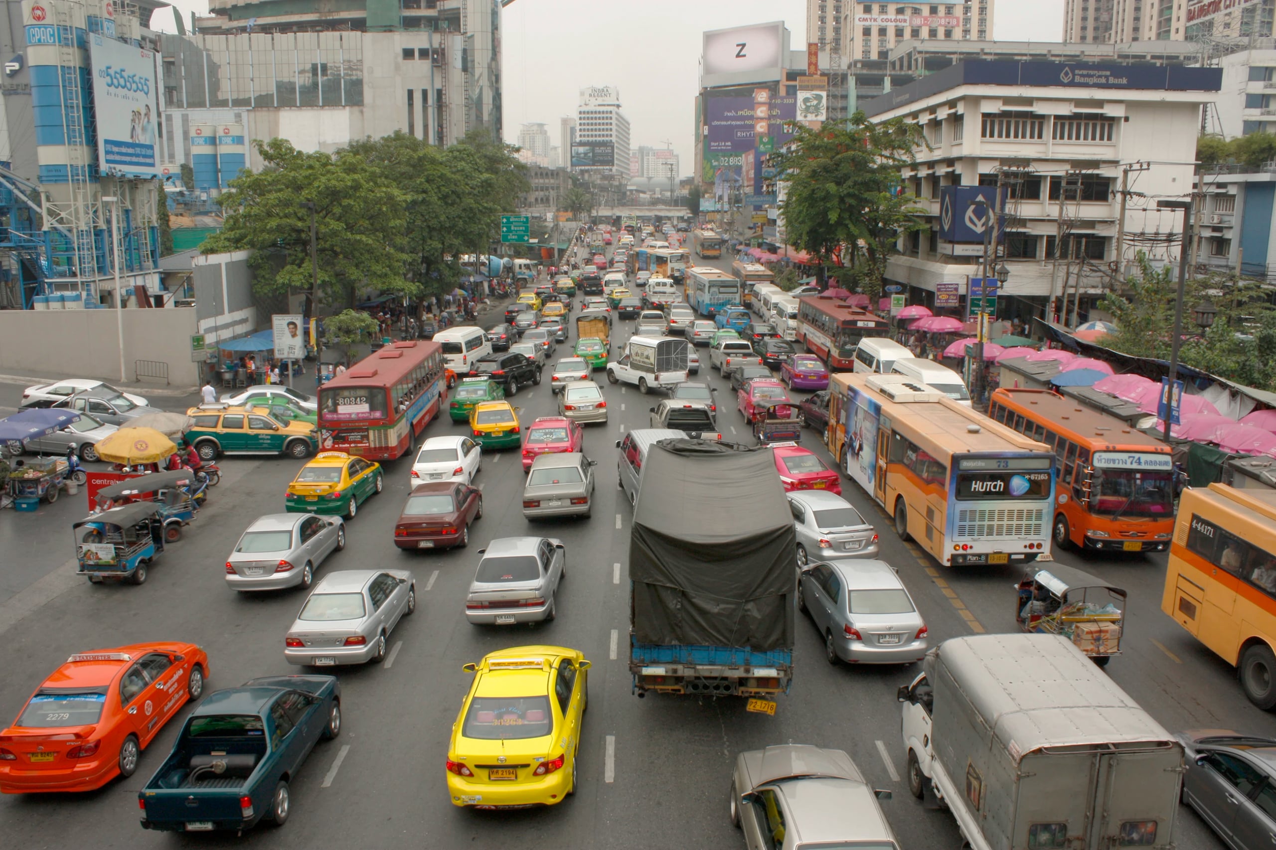 Eight lanes of gridlocked Bangkok traffic seen from above, taxis, tuk-tuks and buses packed between shophouses