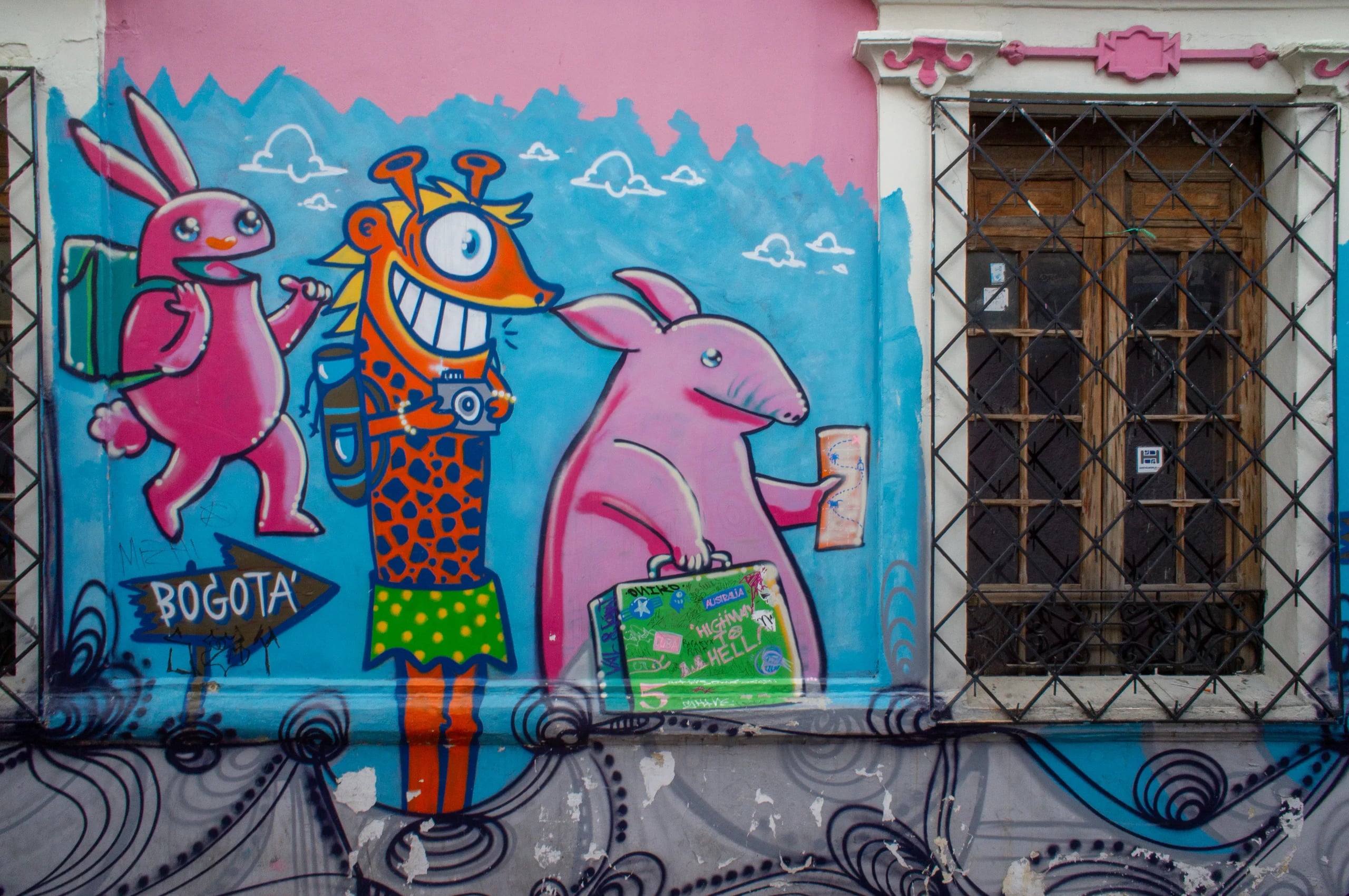 A street mural of a pink rabbit, a grinning giraffe and a pink anteater carrying suitcases beside a signpost reading Bogota