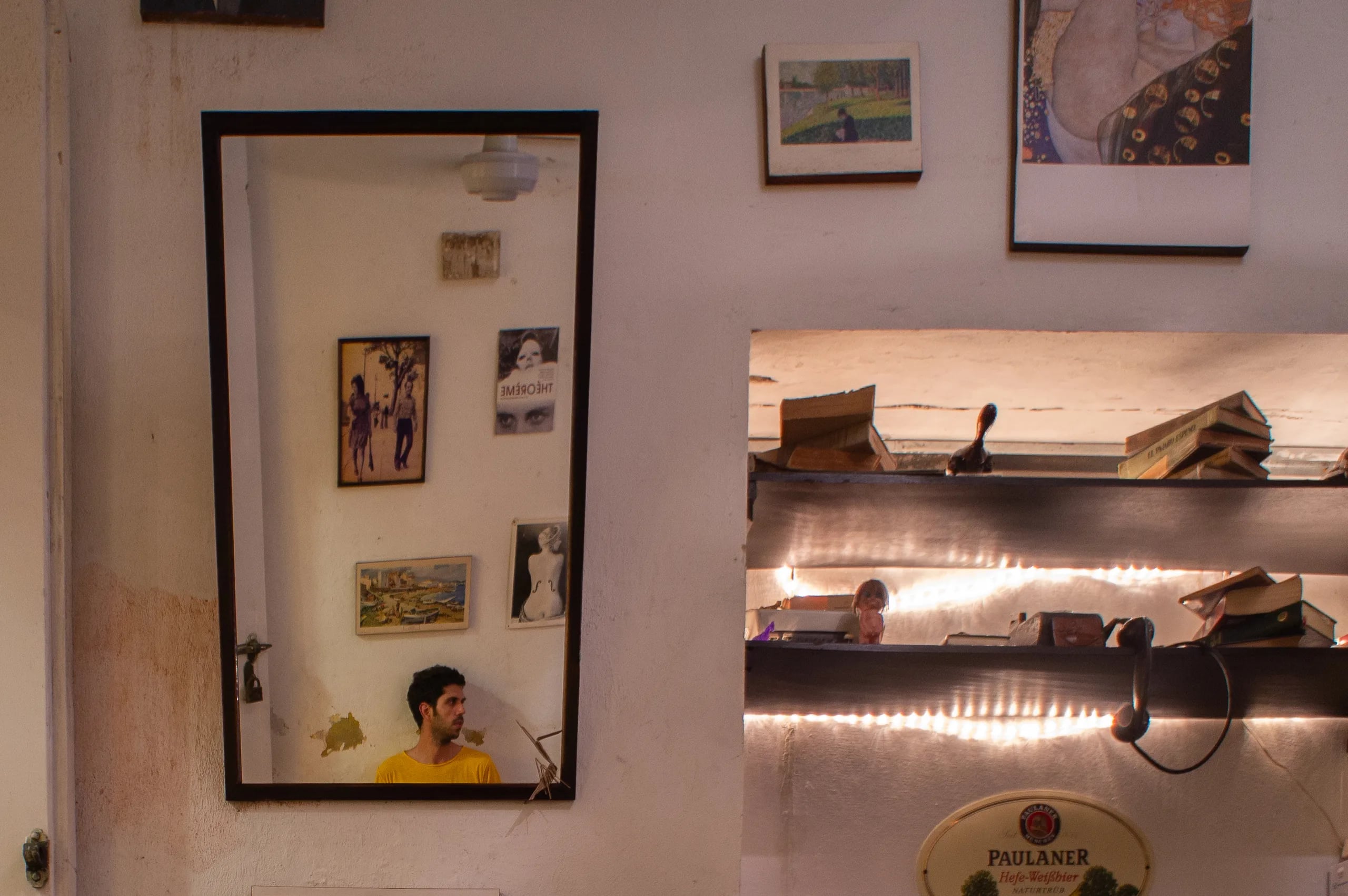 A dim interior wall hung with postcards and prints, a tall mirror reflecting a man in a yellow t-shirt, and a lit shelf of books beside a Paulaner sign.