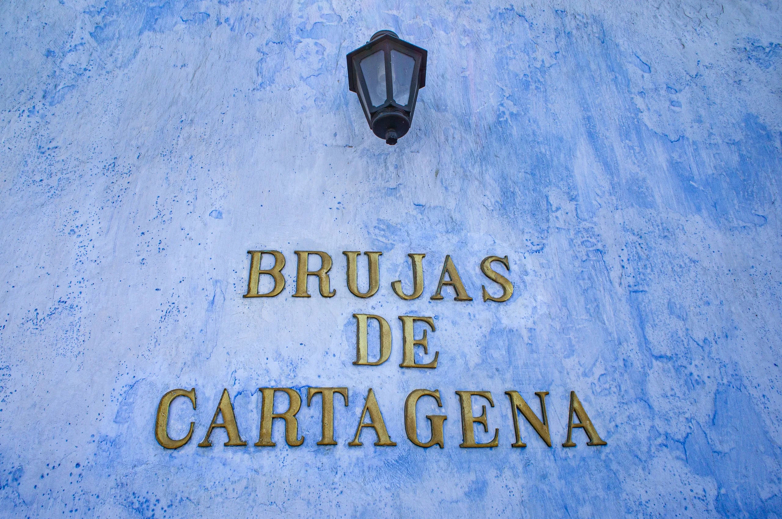 Gold lettering reading “Brujas de Cartagena” fixed to a weathered, mottled blue wall beneath a black lantern.