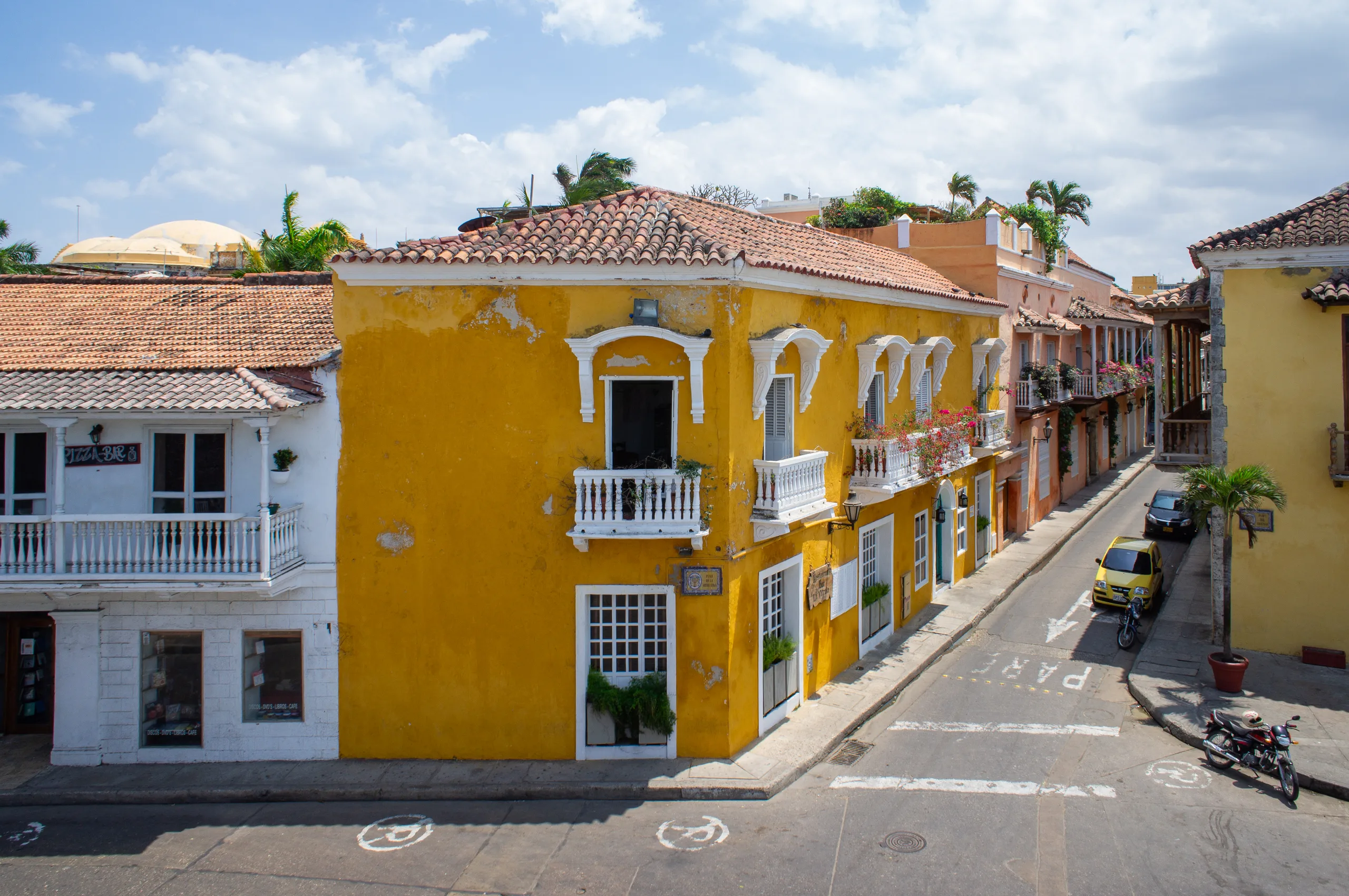 Seen from above, a mustard-yellow corner house with white balconies and red tiled roofs, a yellow taxi and a parked motorbike on the street running away beside it.