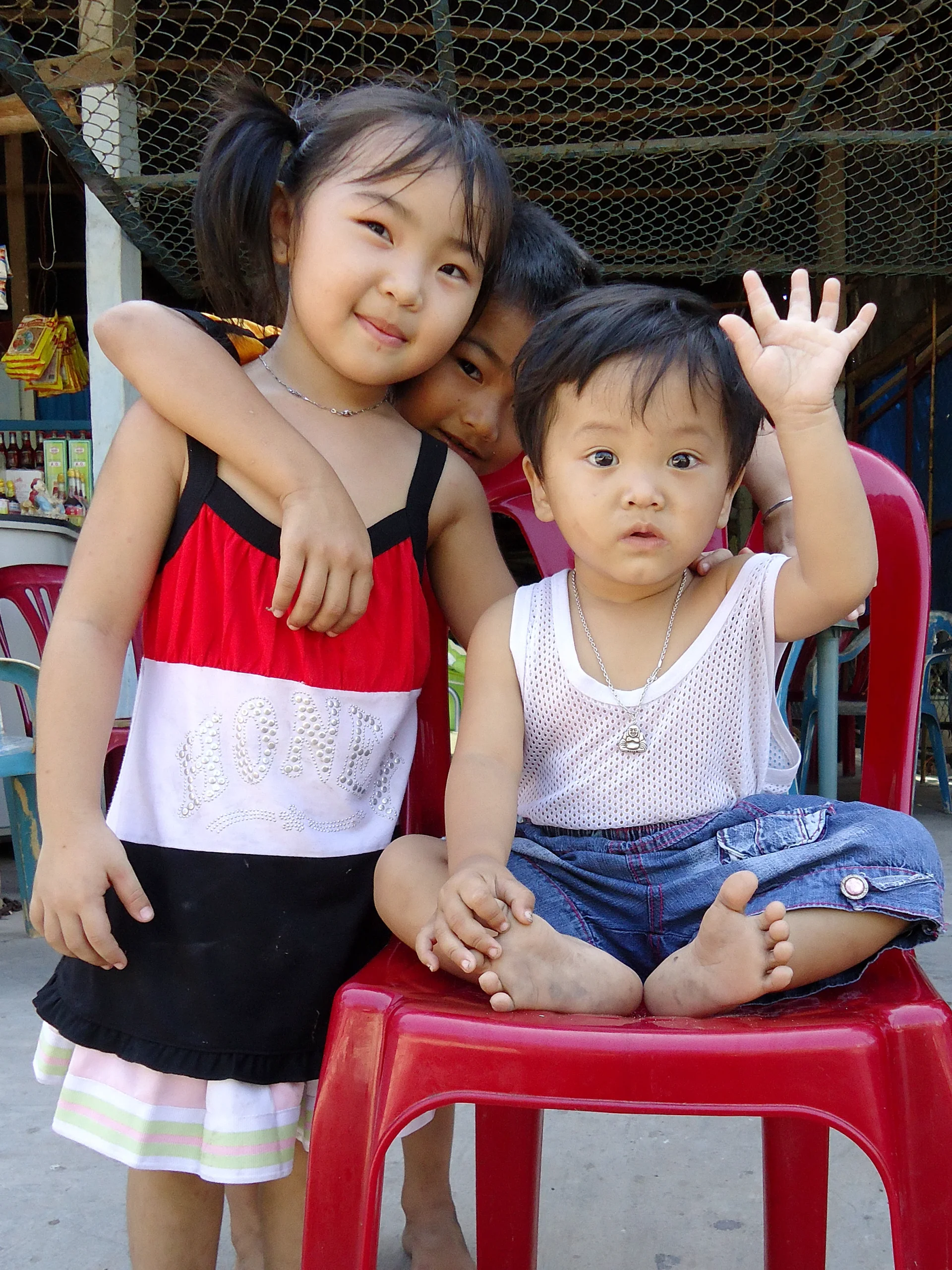 Three small children sharing a red plastic chair, one waving at the camera while the others lean in behind