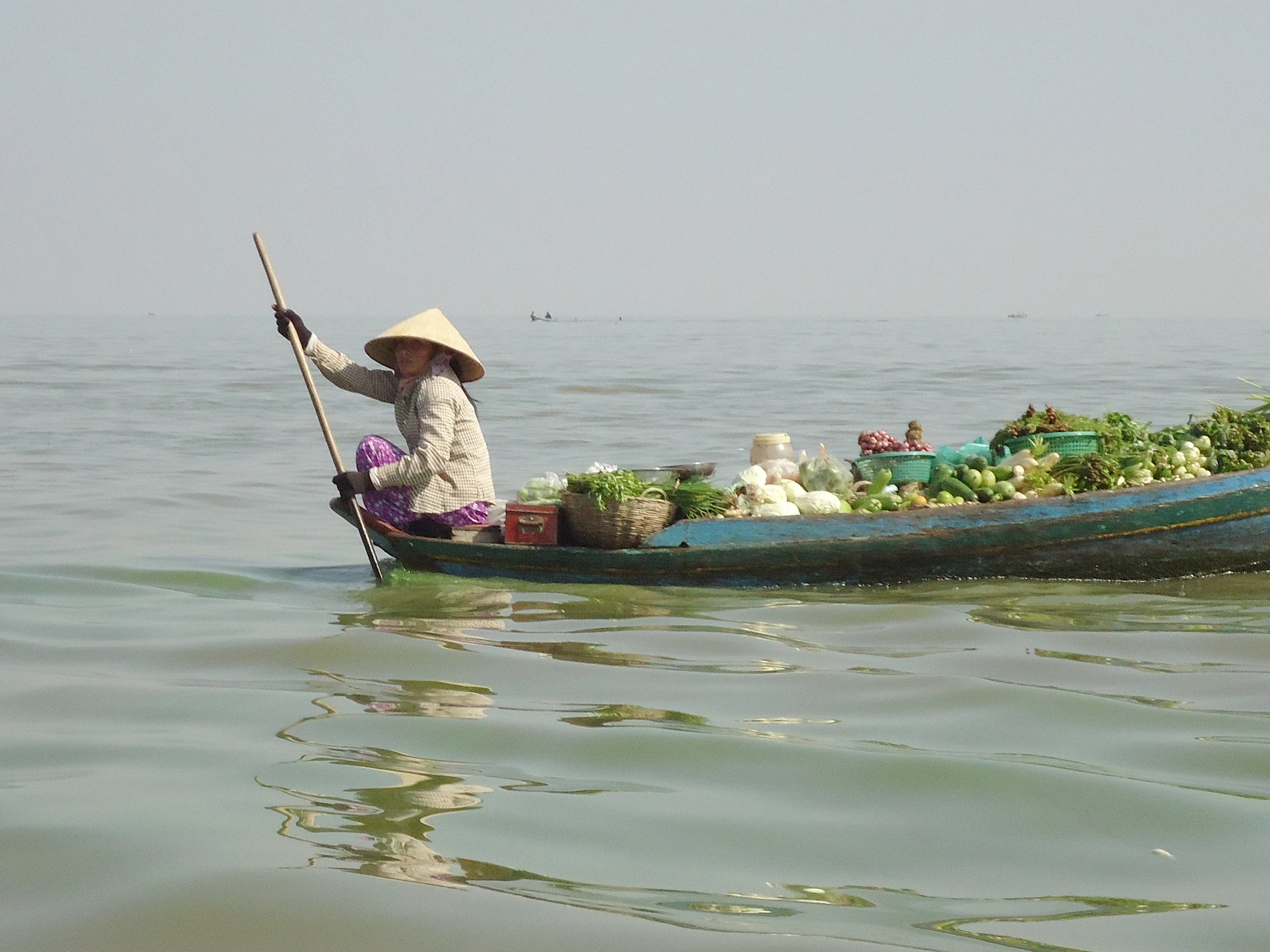 A woman in a conical hat poling a long wooden boat loaded with greens, cabbages and baskets across the lake
