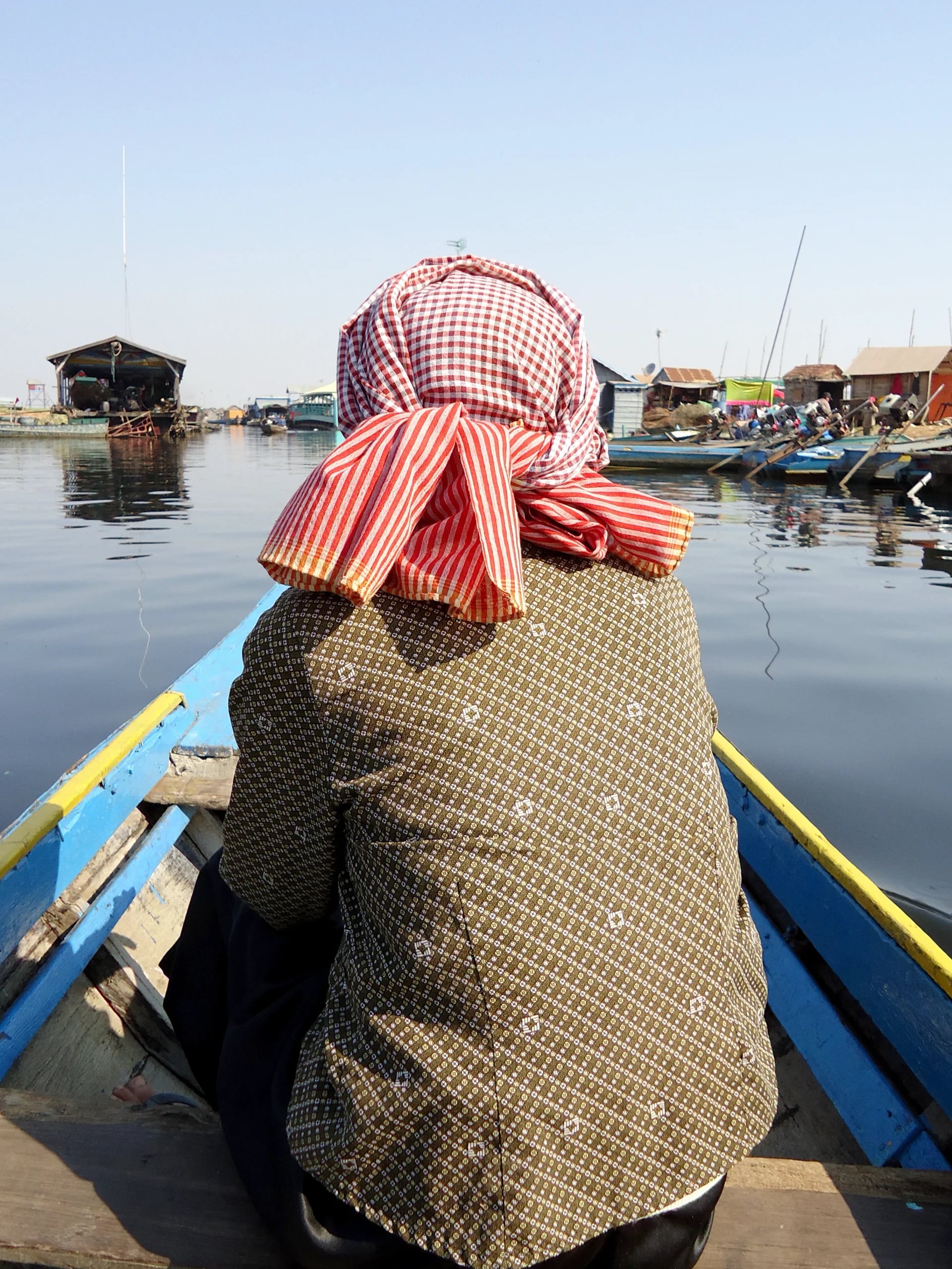 The boatman seen from behind at the tiller, a red-checked krama wrapped round his head, the floating village ahead