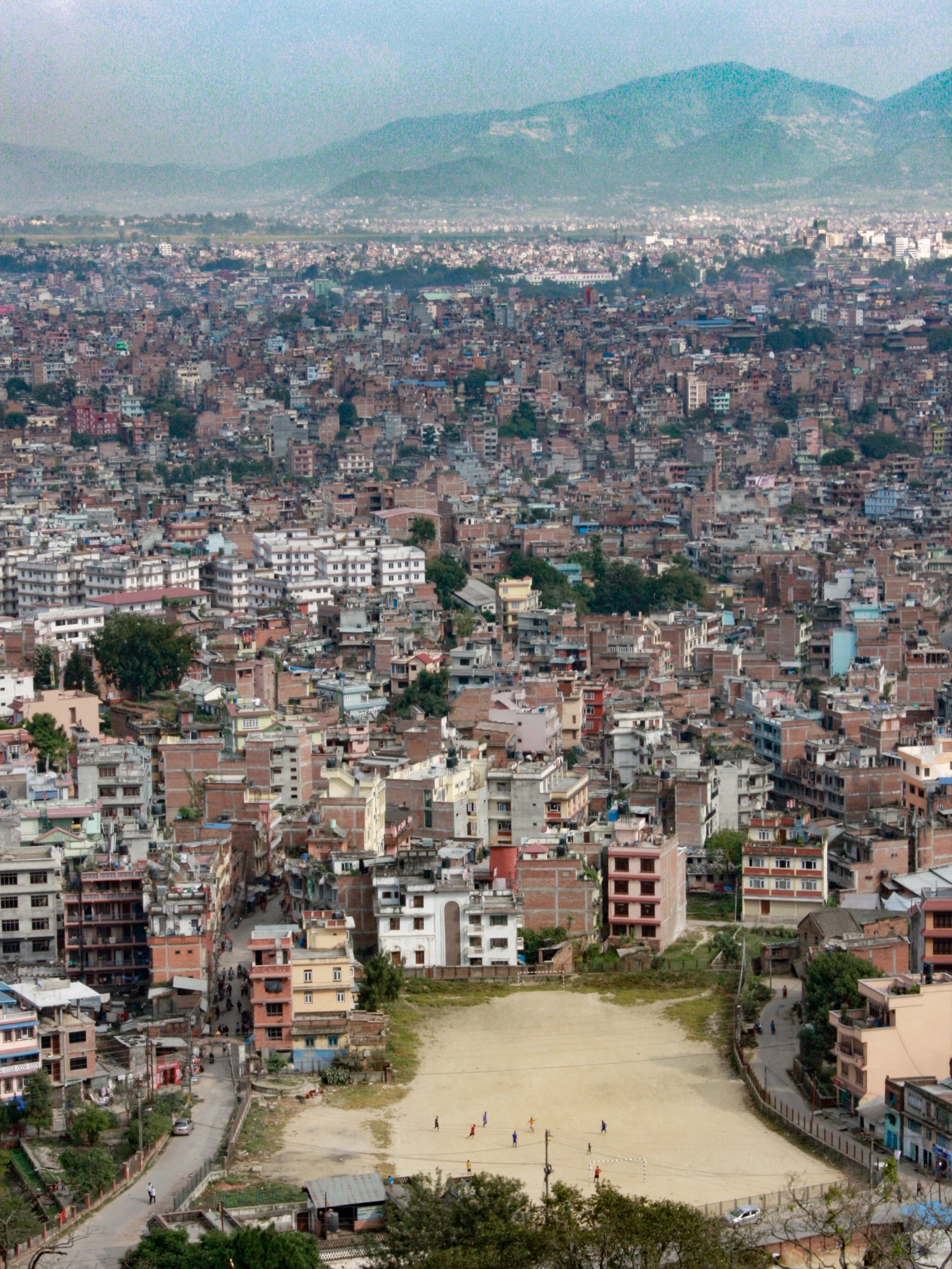 Kathmandu spread out below Swayambhunath, dense rooftops running to the hills with a dusty pitch in the foreground