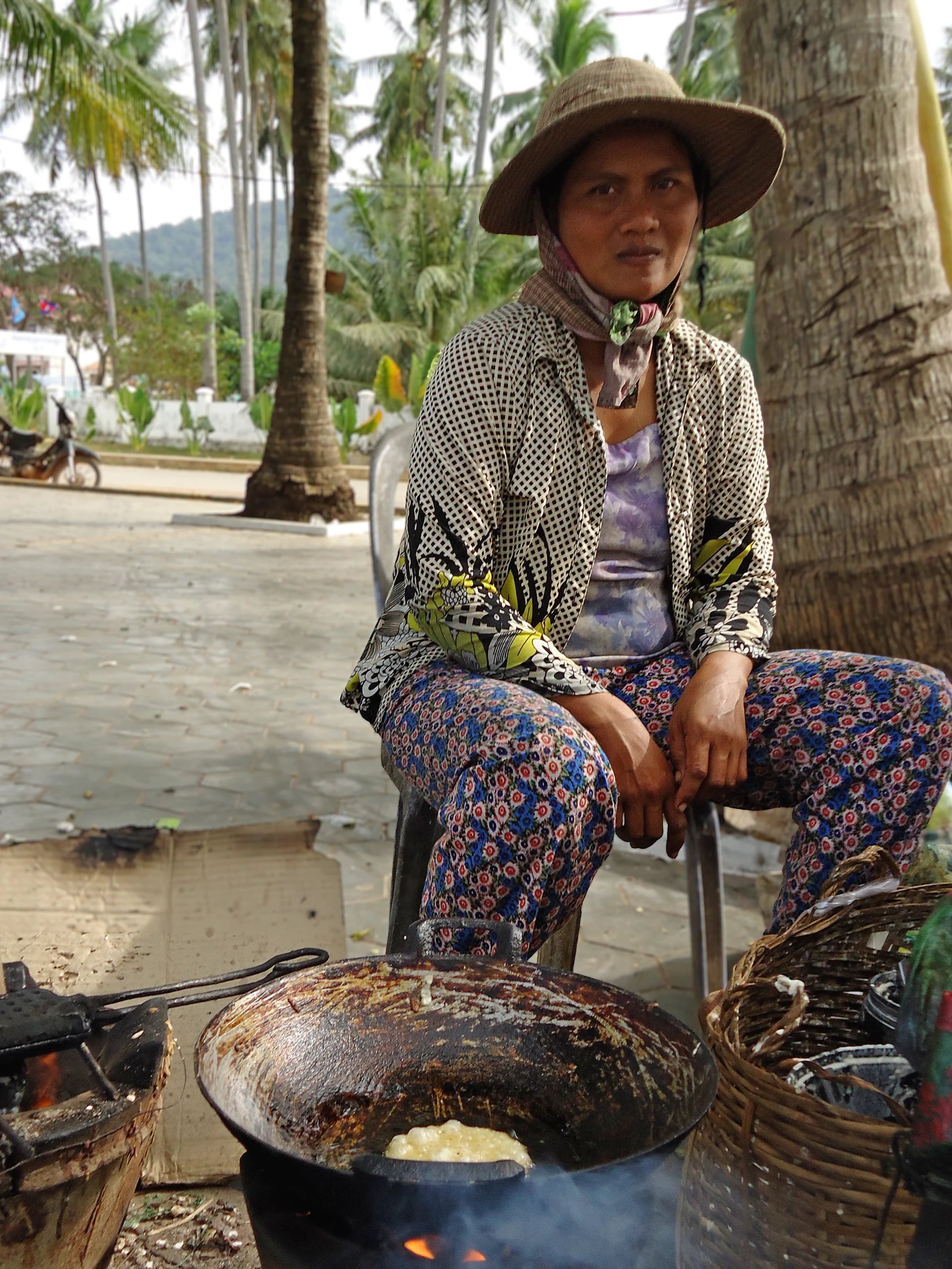 A woman in a straw hat and patterned trousers sitting beside a wok of frying batter over an open fire, palms behind her