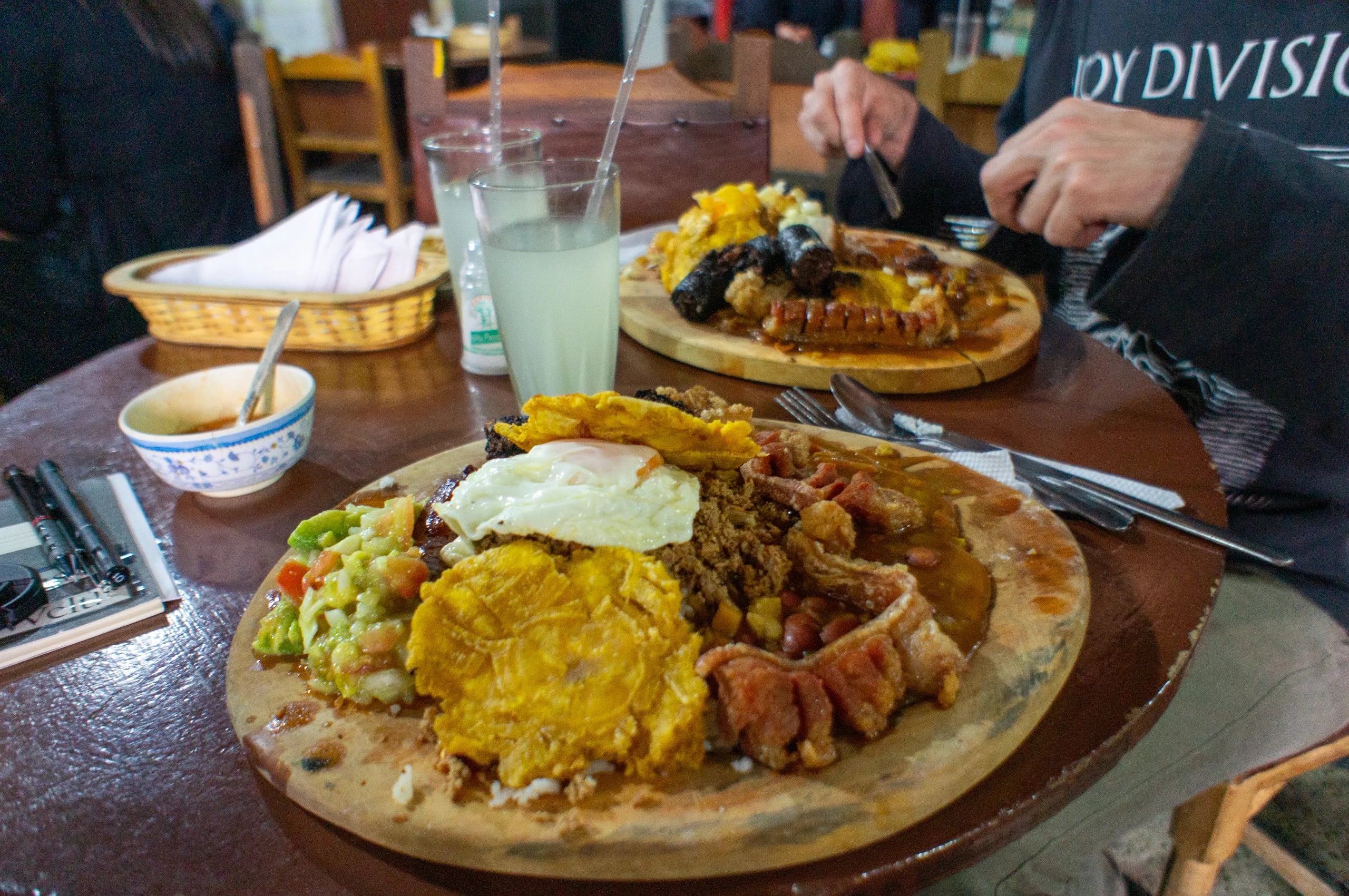 A wooden platter of bandeja paisa on a restaurant table: fried egg, plantain, beans, chicharron, avocado and rice