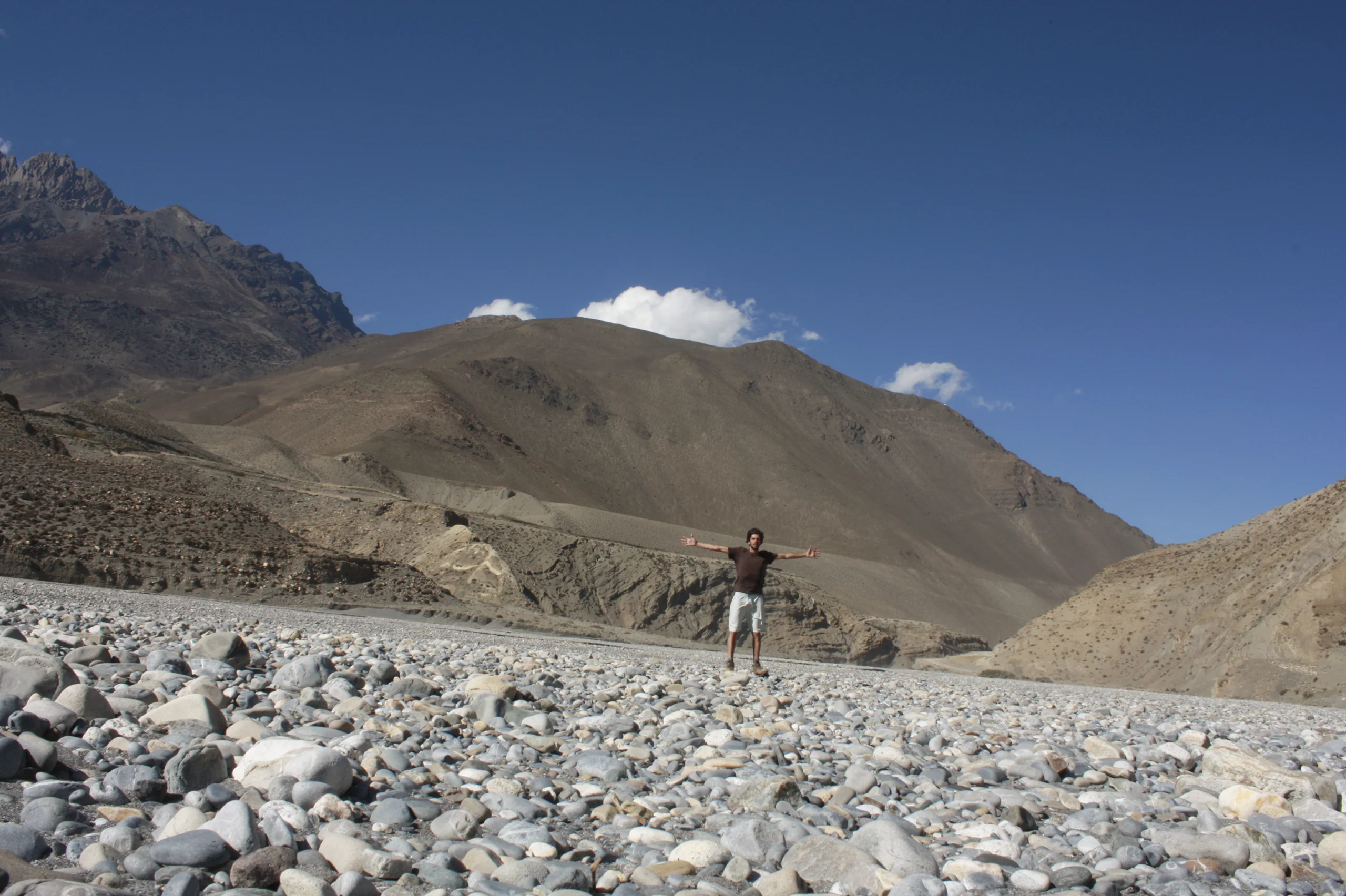 A man in a dark T-shirt and pale shorts stands with his arms flung wide on a broad bed of grey river stones, bare brown mountains behind him.