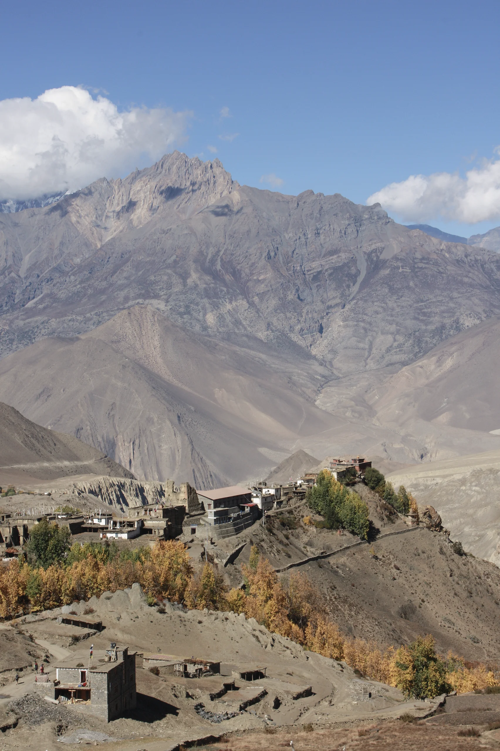 A village of flat-roofed stone houses on a ridge, ringed by poplars turning yellow, beneath an immense grey mountain wall streaked with scree.