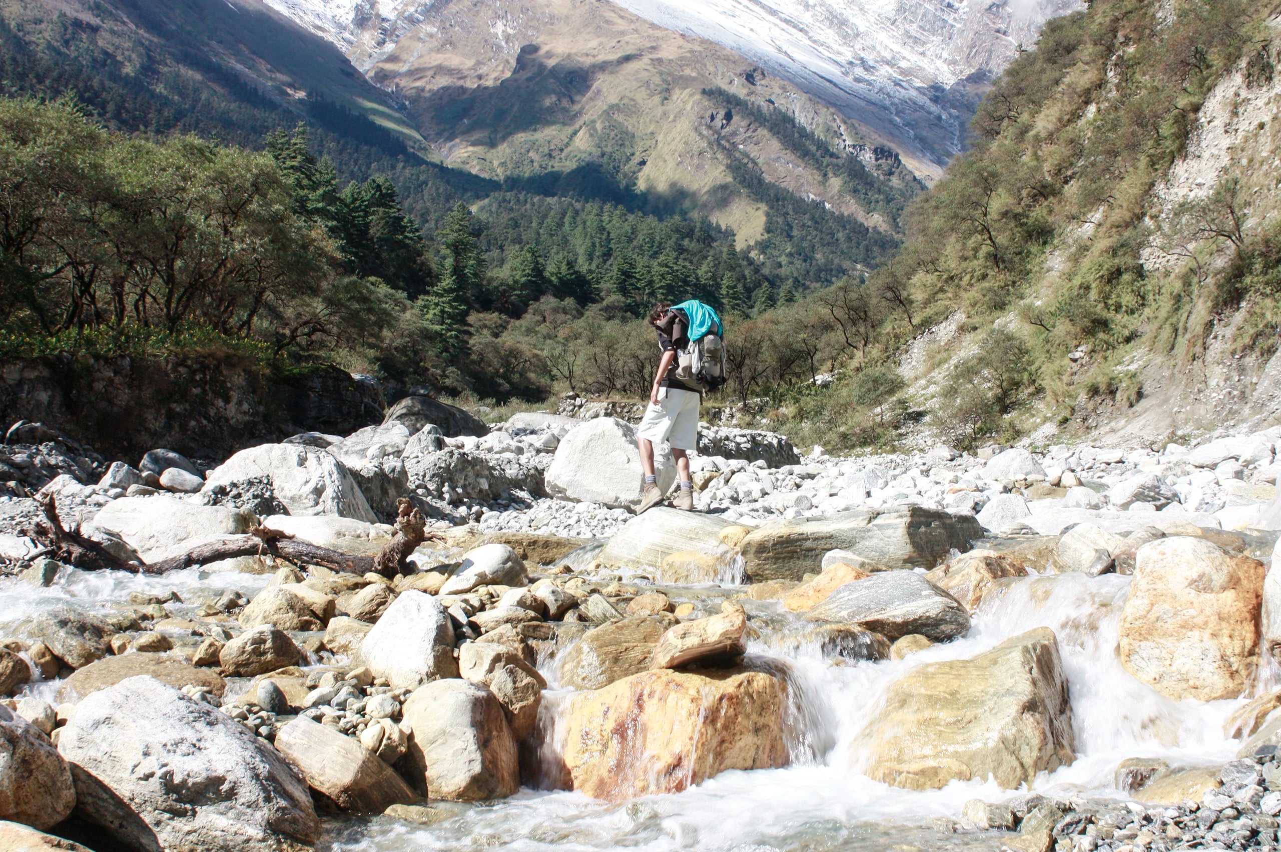 A trekker with a large backpack and a turquoise towel picks his way across pale boulders in a fast, shallow river, snow-dusted peaks closing in at the head of the valley.