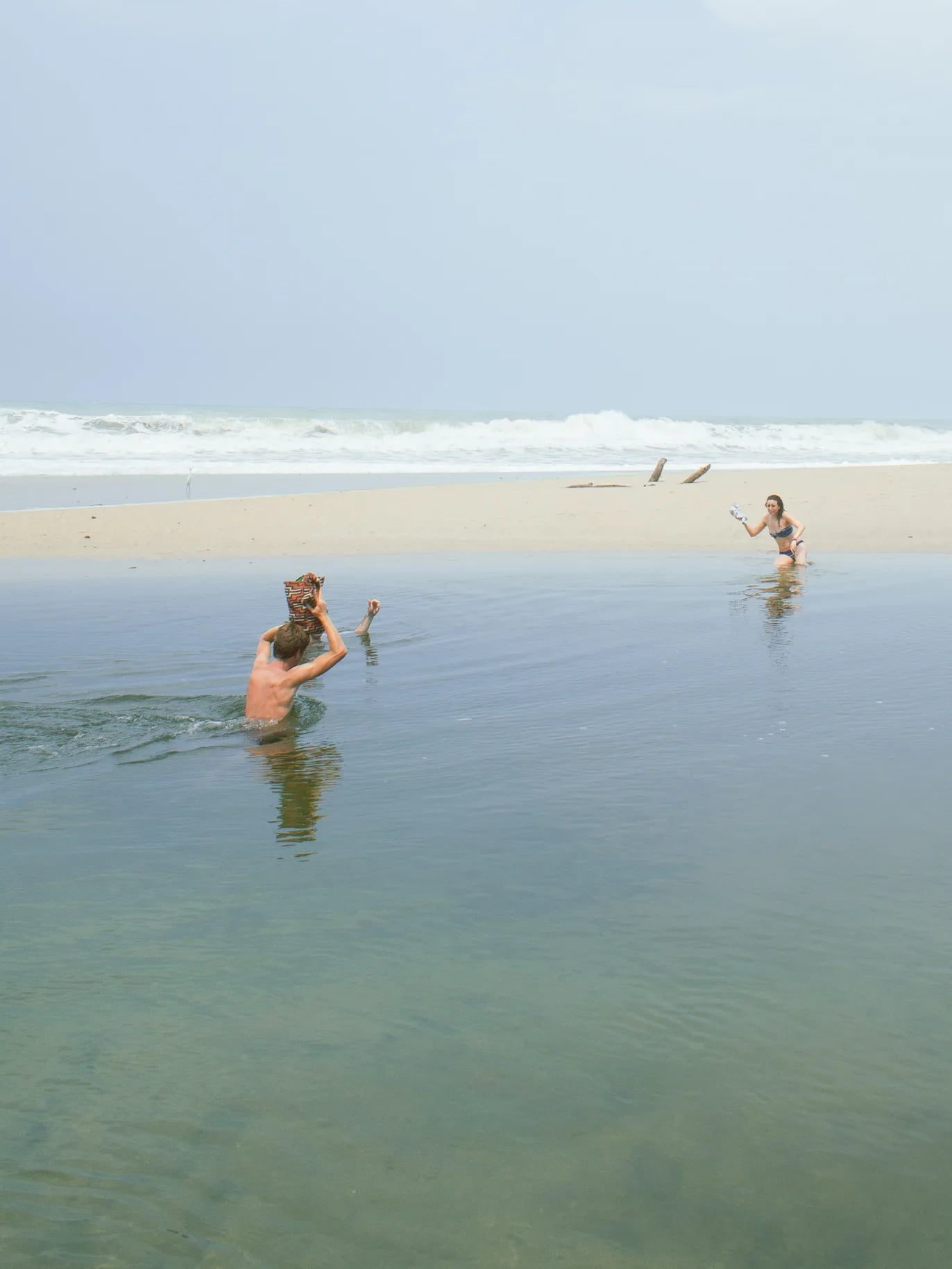Two swimmers in the still water of a river mouth, a sandbar separating them from the surf beyond