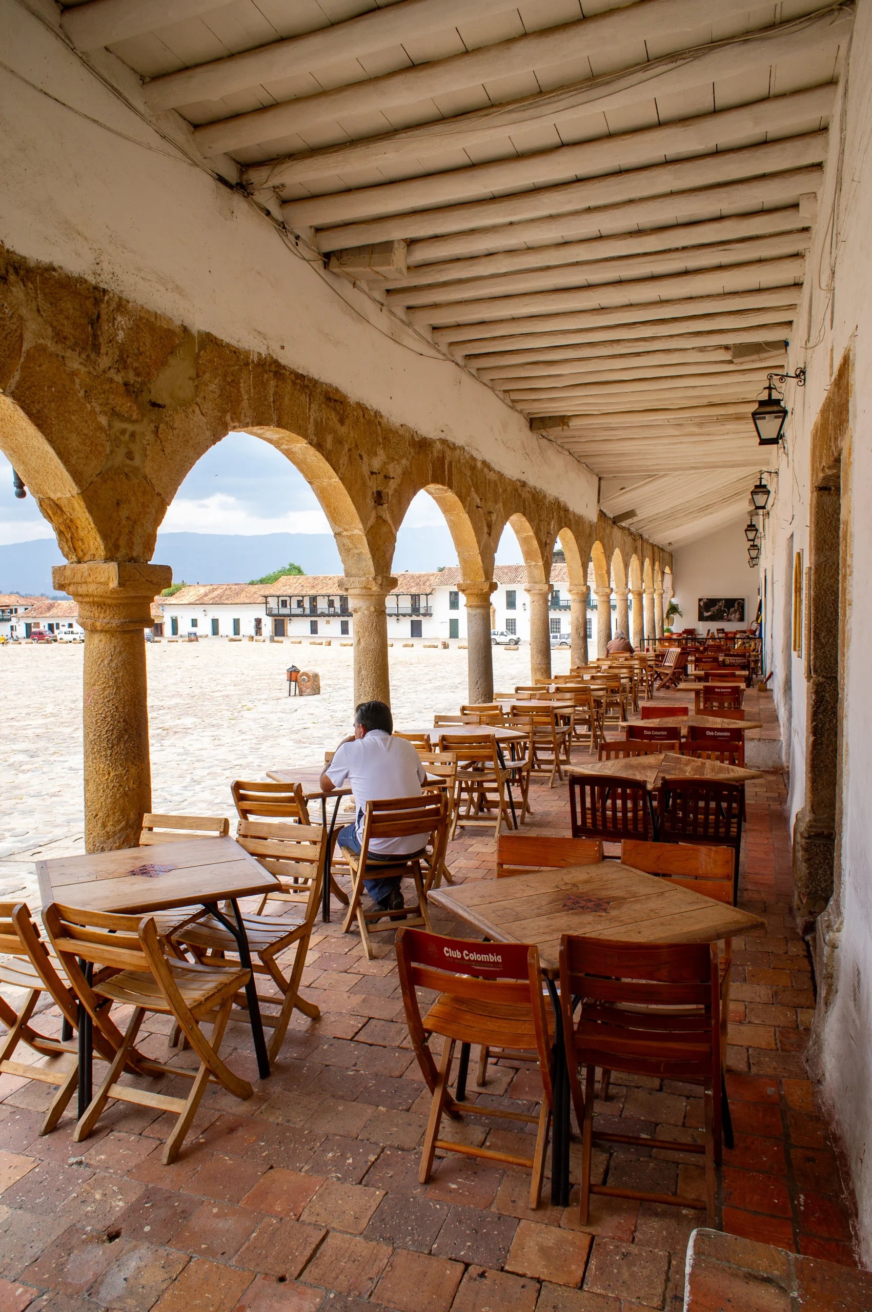 Cafe tables under the stone arcade on Plaza Mayor, the open square and distant hills framed through the arches