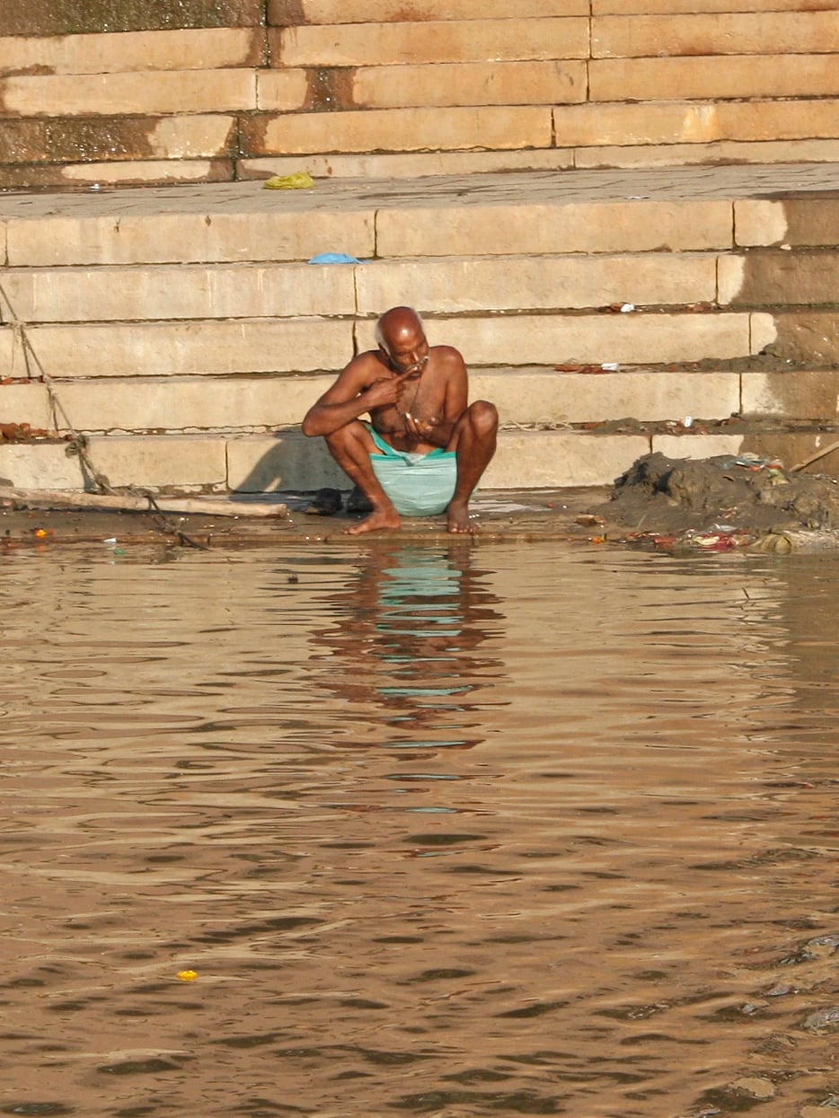 A man crouching at the water's edge on the ghat steps brushing his teeth, his reflection in the Ganges below