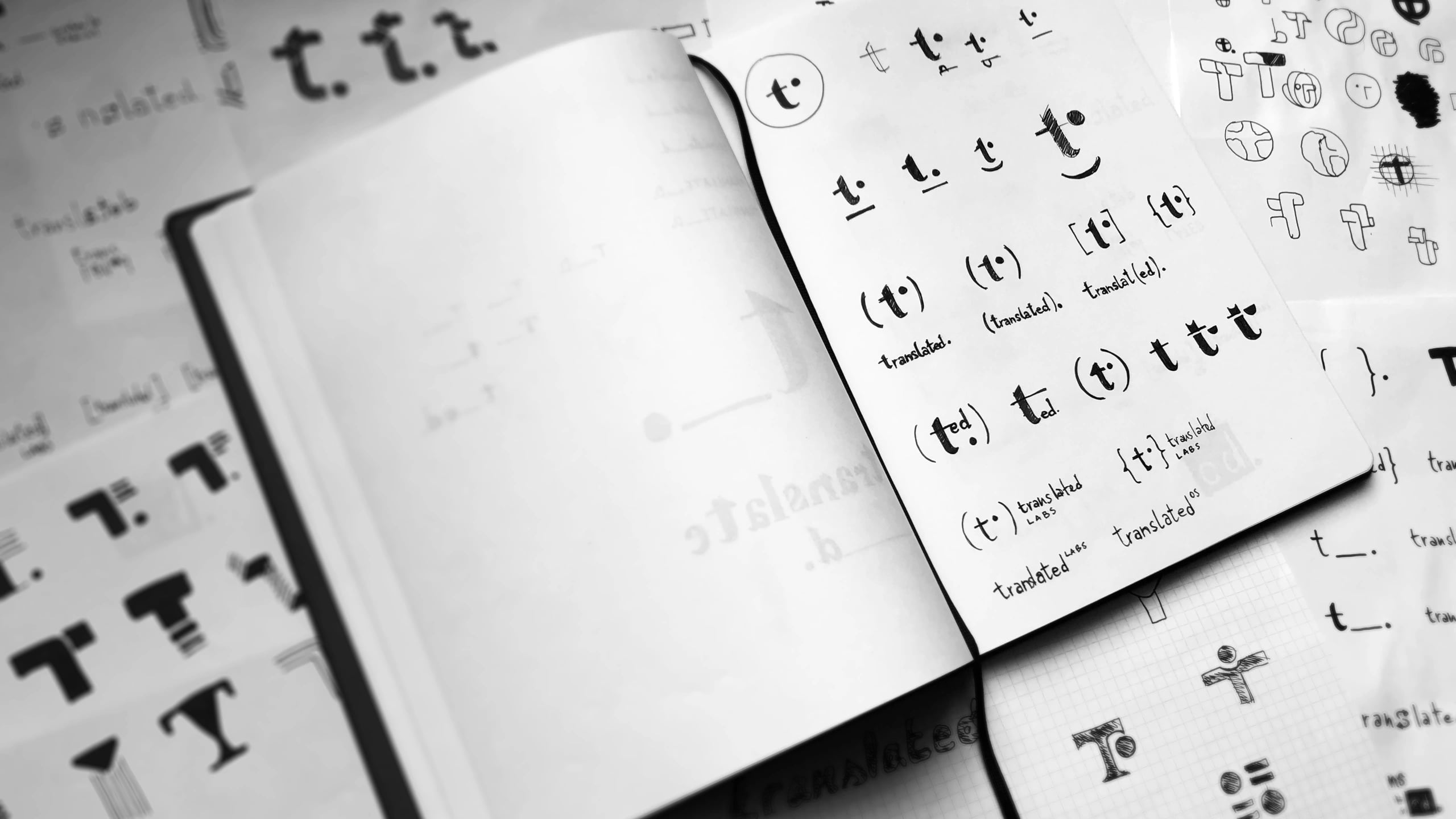 An open sketchbook surrounded by loose sheets of hand-drawn logo studies, all working the lowercase t of translated into a mark