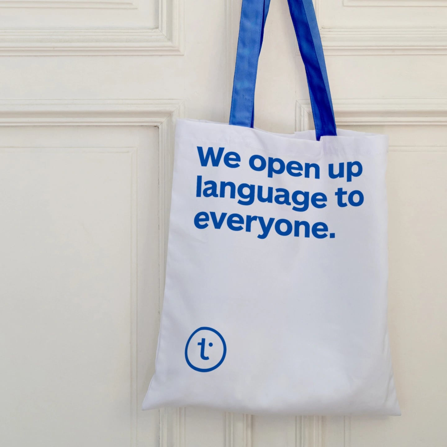 A white tote bag with blue handles hanging on a panelled door, printed with We open up language to everyone above the t mark